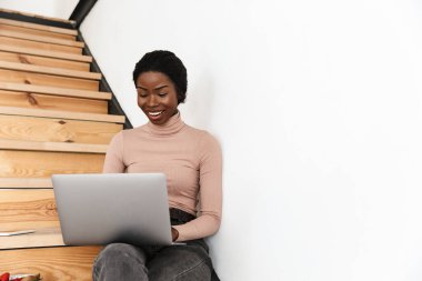 Evdeki merdivenlerde dizüstü bilgisayar kullanan güzel ve iyimser bir Afrikalı kadın resmi