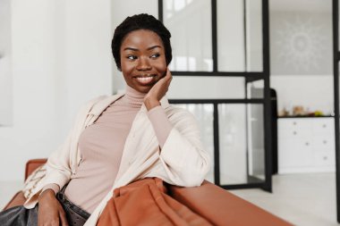 Çekici, gülümseyen Afrikalı bir kadın evdeki koltukta oturuyor, modern iç mekan, rahatlatıcı.