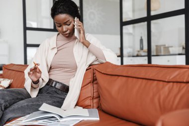 Evdeki koltukta oturan ve cep telefonuyla belgelerin yanında konuşan oldukça ciddi bir Afrikalı kadının fotoğrafı.