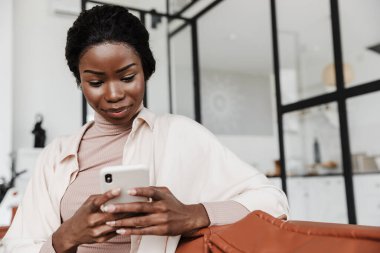Evde cep telefonu kullanırken koltukta oturan inanılmaz konsantre olmuş Afrikalı bir kadının görüntüsü