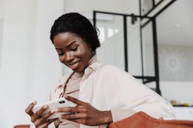 Evdeki kanepede oturup cep telefonuyla video izleyen neşeli, iyimser, genç bir Afrikalı kadının fotoğrafı.