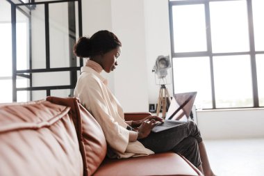 Evde dizüstü bilgisayarla çalışırken kanepede oturan Afrikalı güzel bir kadının yan görüntüsü.