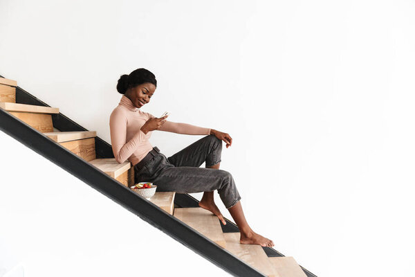 Image of beautiful happy cheerful cute young african woman sitting on stairs indoors at home while using mobile phone