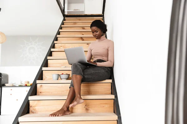ラップトップコンピュータを使用しながら家の中で階段の上に座って美しい濃縮若いアフリカの女性のイメージ — ストック写真
