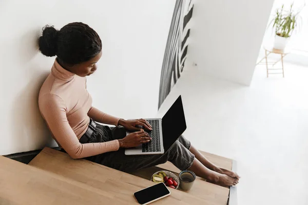 一个有魅力的年轻非洲女人坐在家里的楼梯上 在笔记本电脑上工作 吃早餐 — 图库照片