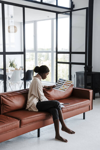 Photo of young african concentrated woman interior designer sitting on sofa indoors at home while working with catalogue of colors
