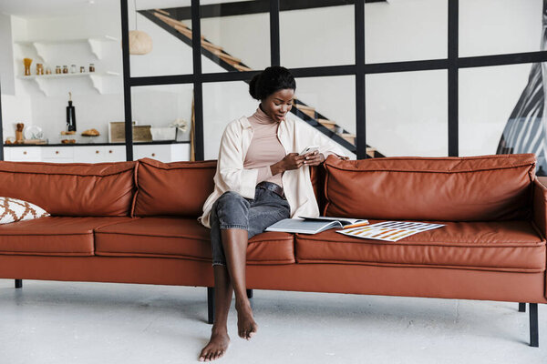 Picture of young concentrated woman interior designer sitting on sofa indoors at home while working with catalogue of colors and using mobile phone
