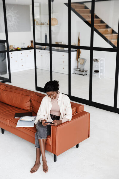 Image of amazing cheery young african woman sitting on sofa indoors at home while using mobile phone