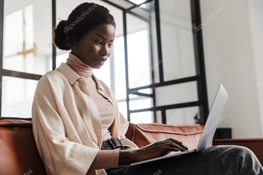 Photo of young concentrated woman sitting on sofa indoors at home while working with laptop computer