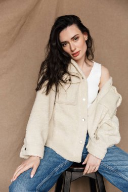 Çekici ceketli genç bir kadının bej arka planda izole edilmiş bir sandalyede otururken kameraya baktığı fotoğraf.