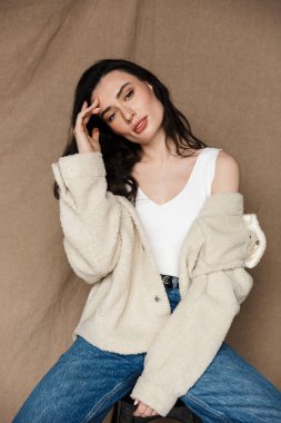 Çekici ceketli genç bir kadının bej arka planda izole edilmiş bir sandalyede otururken kameraya baktığı fotoğraf.