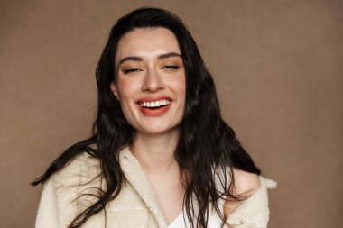 Ceketli neşeli genç bir kadının bej arka planda izole edilmiş kameraya bakıp güldüğü fotoğraf.