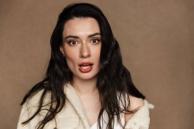 Ceketli genç bir kadının bej arka planda izole edilmiş kameraya şaşırdığını gösteren bir fotoğraf.