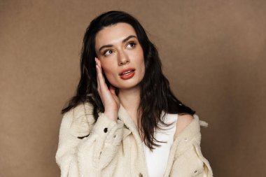 Ceketli genç bir kadının bej arka planda izole edilmiş bir şekilde poz vermesi ve bakması.