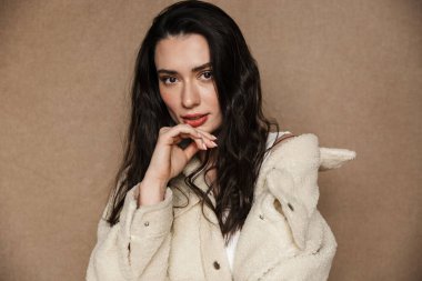 Çekici, ceketli, poz veren ve bej arka planda izole edilmiş kameraya bakan hoş bir kadının fotoğrafı.