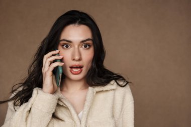 Ceketli, güzel, esmer bir kadının bej arka planda cep telefonuyla konuşurken çekilmiş fotoğrafı.
