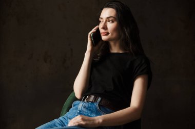 Sandalyede oturan ve cep telefonuyla konuşan güzel bir genç kadının fotoğrafı karanlık arka planda izole edilmiş.