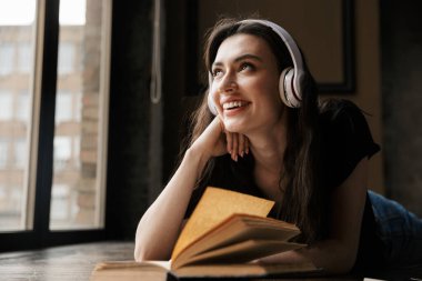 Kulaklık takmış güzel esmer bir kadının ahşap zeminde kitapla yattığı bir fotoğraf.