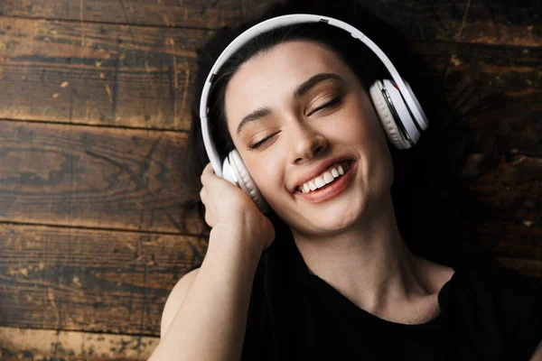 写真上から美しい若いブルネットの女性のヘッドフォンを身に着けている笑顔と木製の床に横たわっ — ストック写真