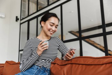 Çekici, gülümseyen Asyalı bir kadın. Günlük kıyafetler giyip evdeki deri koltukta rahatlıyor. Cep telefonu kullanıyor.
