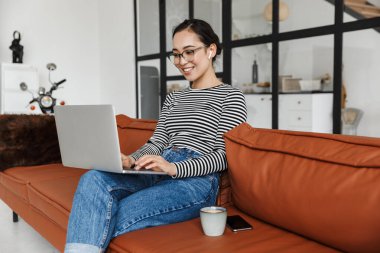 Çekici, gülümseyen Asyalı bir kadın. Günlük kıyafetler giyip evdeki deri koltukta rahatlıyor. Dizüstü bilgisayarla çalışıyor.
