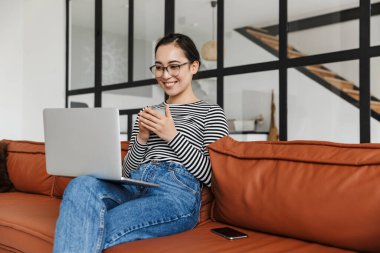 Çekici, gülümseyen Asyalı bir kadın. Günlük kıyafetler giyip evdeki deri koltukta dinleniyor. Video çağrısı yapıyor.