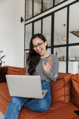 Çekici gülümseyen Asyalı genç iş kadını evdeki deri koltukta dinleniyor, bilgisayardaki video görüşmesi sırasında, kulaklık takıyor, el sallıyor.