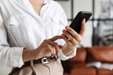 Modern bir oturma odasında cep telefonu kullanan, beyaz tişörtlü, Asyalı genç bir kadının fotoğrafını çek.