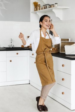 Mutfakta kablosuz kulaklıkla müzik dinlerken önlük giyen neşeli Asyalı kadın.