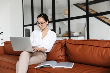 Çekici gülümseyen Asyalı iş kadını evde deri koltukta dinleniyor, dizüstü bilgisayarla çalışıyor.