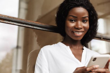 Çekici, gülümseyen, zeki, Afrikalı bir kadın girişimci camdan bir duvara yaslanmış, cep telefonu kullanıyor.