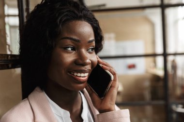 Çekici, gülümseyen, zeki, Afrikalı, genç bir girişimci camdan duvara yaslanmış, cep telefonuyla konuşuyor.