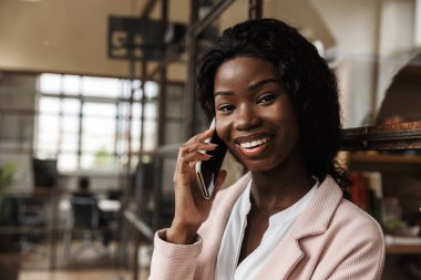 Çekici, gülümseyen, zeki, Afrikalı, genç bir girişimci camdan duvara yaslanmış, cep telefonuyla konuşuyor.