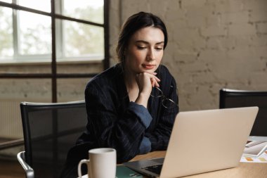 Ofis masasında otururken dizüstü bilgisayarda çalışan kendine güvenen çekici genç bir girişimci.