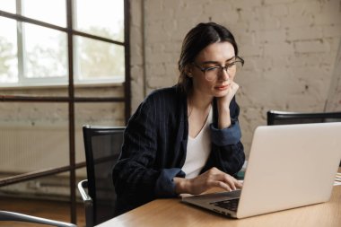 Ofis masasında otururken dizüstü bilgisayarda çalışan kendine güvenen genç ve çekici bir girişimci.