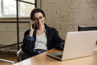 Ofis masasında oturup cep telefonuyla konuşurken dizüstü bilgisayarda çalışan kendine güvenen genç girişimci.