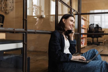 Ofiste oturan, camdan duvara yaslanan, dizüstü bilgisayar kullanan, cep telefonuyla konuşan çekici, genç bir kadın.
