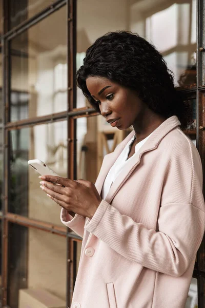 携帯電話を使ってガラスの壁にもたれている間に立っている魅力的なスマートアフリカの若い女性起業家 — ストック写真