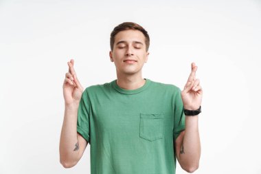 Beyaz arka planda dua eden ve parmaklarını çapraz yapan beyaz tişörtlü çekici beyaz adam.