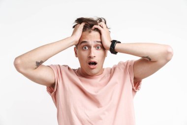 Temel tişörtlü korkmuş beyaz adamın beyaz arka planda izole edilmiş bir şekilde stres altında ve başını tutarken görüntüsü.