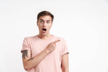 Temel tişörtlü, beyaz arka planda izole edilmiş, merak ve işaret eden şok olmuş genç adam görüntüsü.
