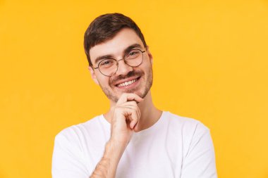 Gözlüklü neşeli bir gencin fotoğrafı. Kameraya bakıyor ve sarı arka planda izole bir şekilde gülümsüyor.