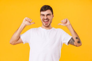 Gözlüklü heyecanlı genç adamın sarı arka planda izole edilmiş parmaklarını gösteren fotoğrafı.