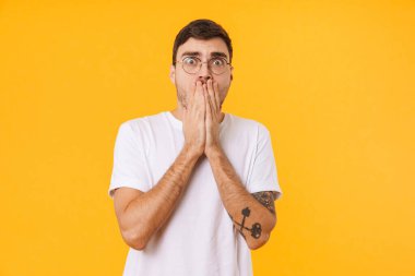 Gözlüklü korkmuş bir gencin fotoğrafı. Kameraya bakıyor ve sarı arka planda ağzını kapatıyor.