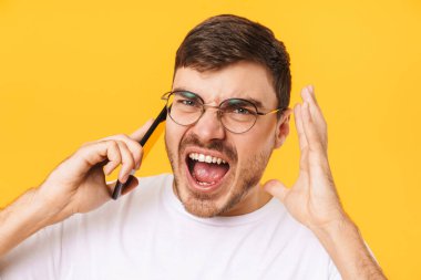 Gözlüklü kızgın beyaz adamın fotoğrafı. Akıllı telefondan konuşuyor ve sarı arka planda izole bir şekilde çığlık atıyor.