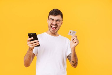 Gözlüklü, elinde kredi kartı ve cep telefonu olan mutlu bir gencin fotoğrafı sarı arka planda izole edilmiş.