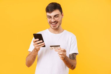 Gözlüklü, elinde kredi kartı ve cep telefonu olan mutlu bir gencin fotoğrafı sarı arka planda izole edilmiş.