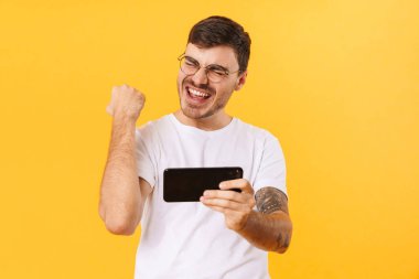 Sarı arka planda izole edilmiş cep telefonuyla video oyunu oynayan gözlüklü heyecanlı kıllı adamın fotoğrafı.