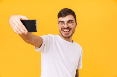Gözlüklü neşeli genç adamın cep telefonuyla selfie çektiği ve sarı arka planda izole bir şekilde gülümsediği fotoğraf.