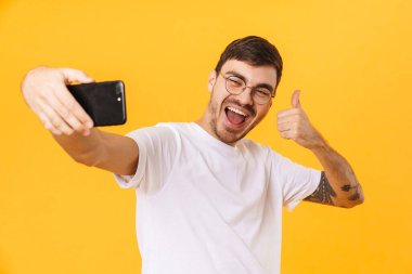 Gözlüklü neşeli bir gencin cep telefonuyla selfie çektiği ve sarı arka planda izole bir şekilde baş parmağını gösterdiği fotoğraf.